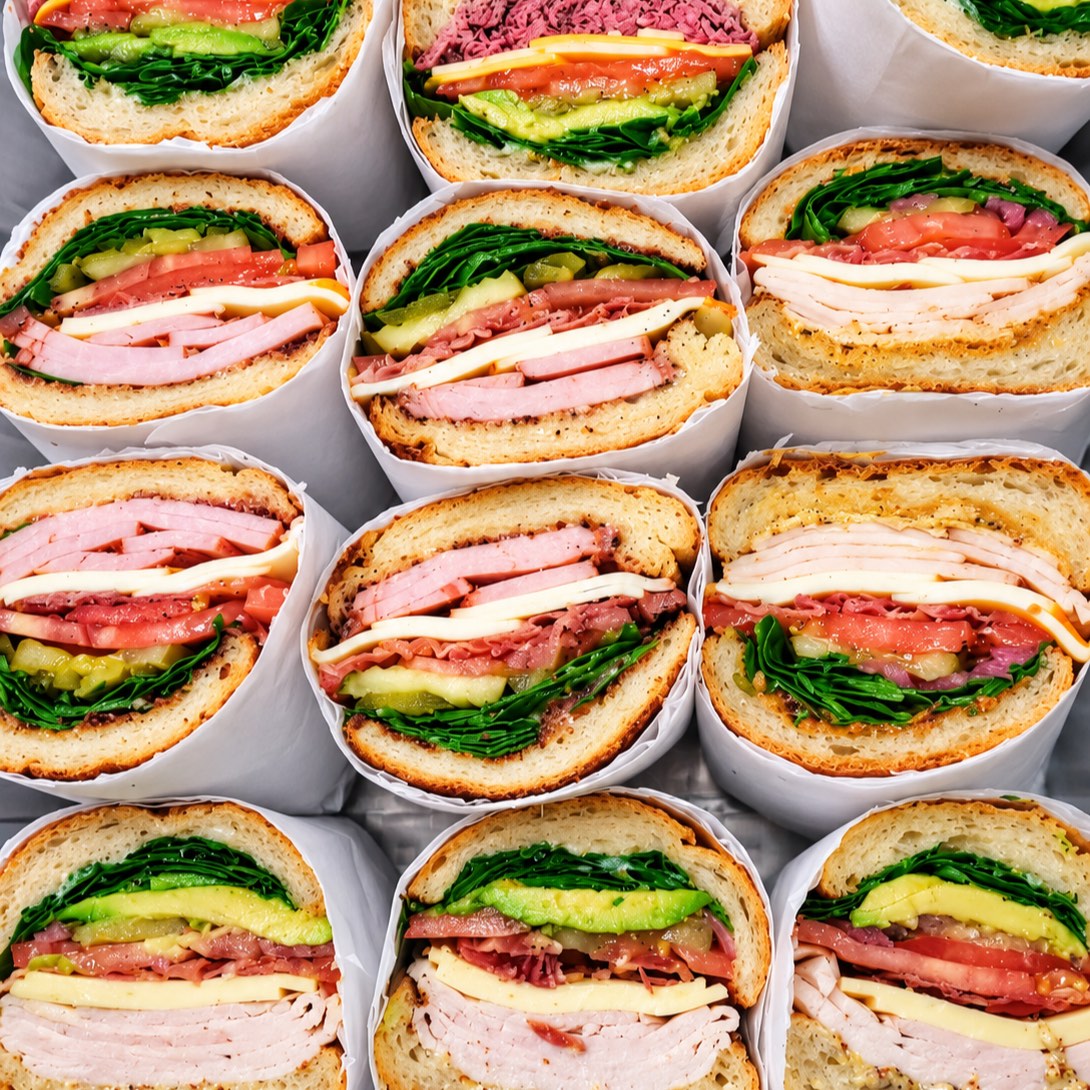 Rhea’s sandwiches cut in half, wrapped in white paper and packed in rows: ham, turkey, cheese, tomato, avocado, pickles and greens on toasted rolls