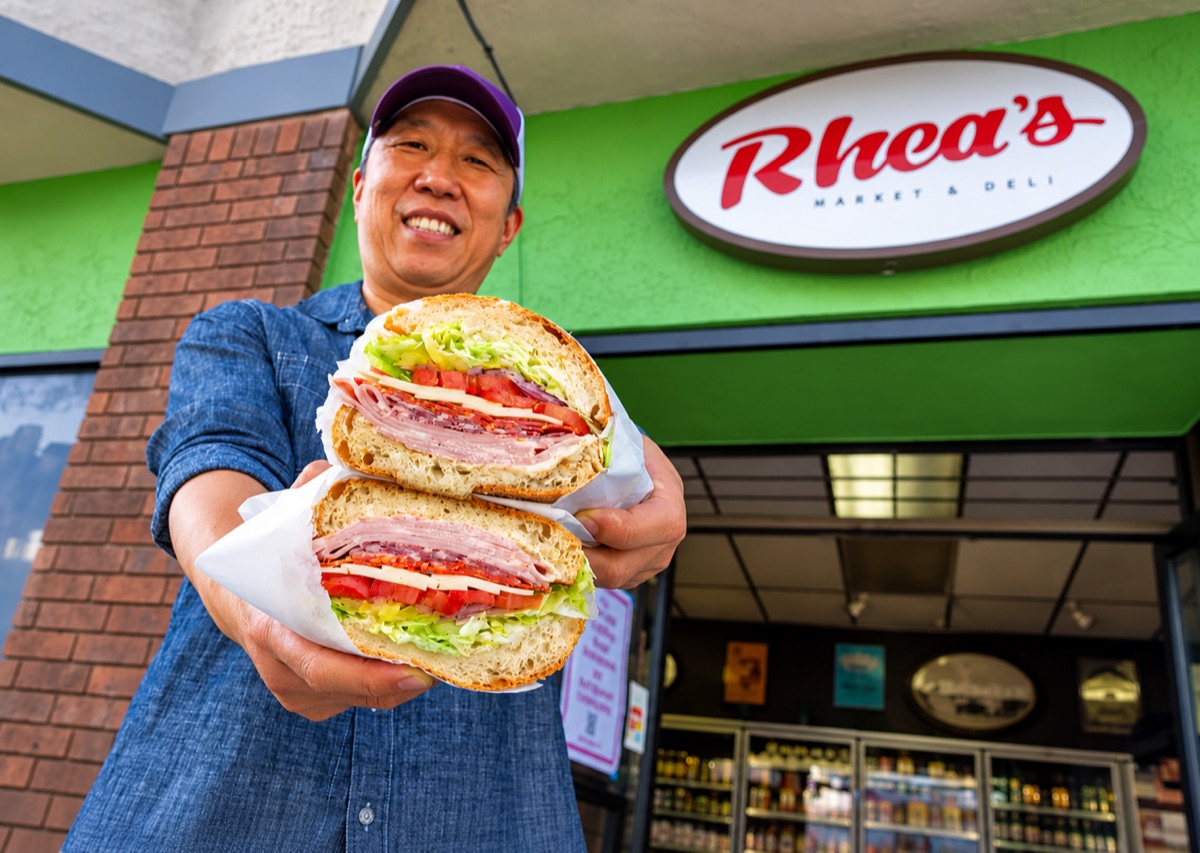 Rhea’s founder smiling outside the shop under the oval Rhea’s Market & Deli sign, holding a sandwich cut in half out toward the camera